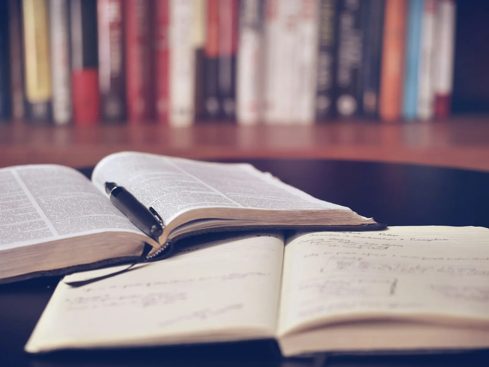 Person sitting and reading an open book, with a notebook and a cup of coffee on a wooden table nearby.