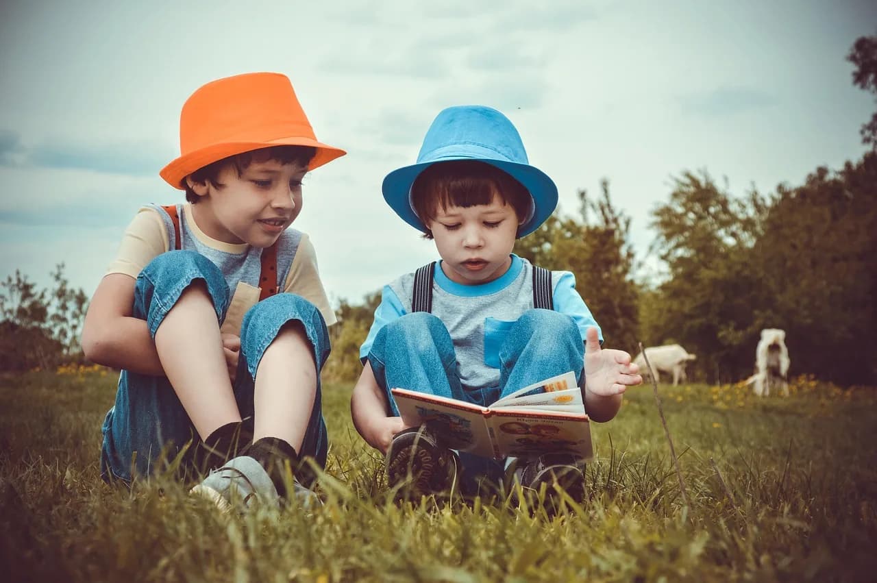 Two boys sitting outdoors in a meadow reading an illustrated storybook together.