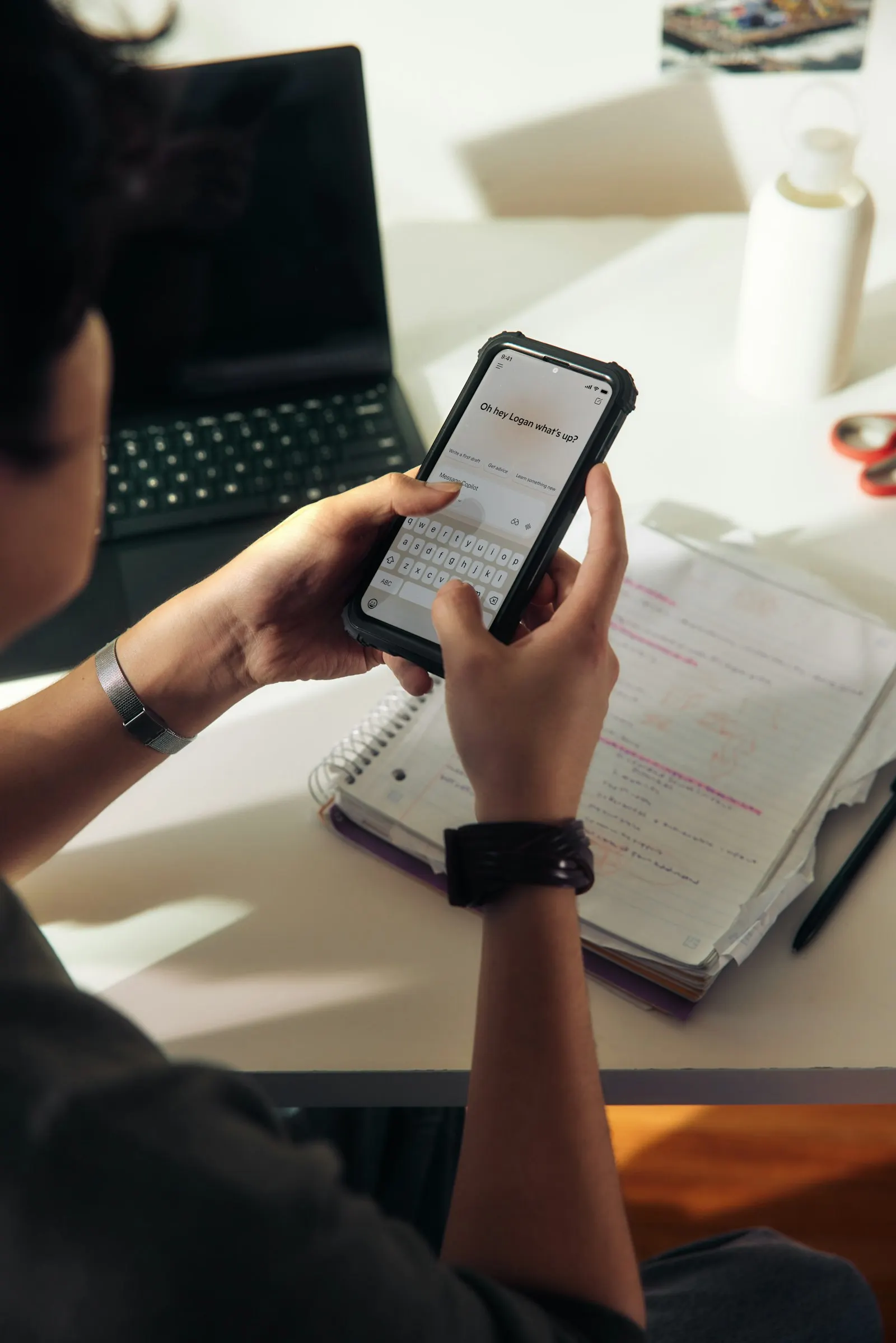 A person checks a learning app on their phone, sitting at a desk with an open notebook.